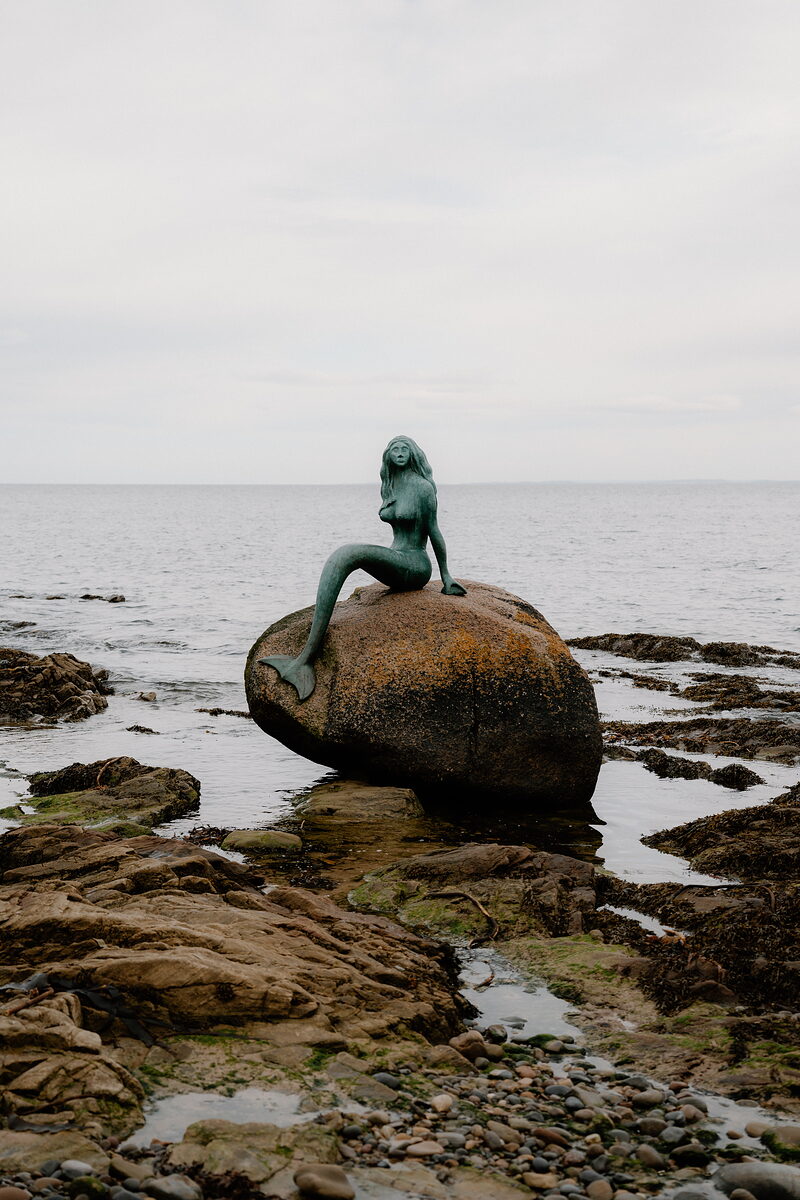 A bronze mermaid statue sits atop a boulder off the Ross and Cromarty shore, framed by calm sea.