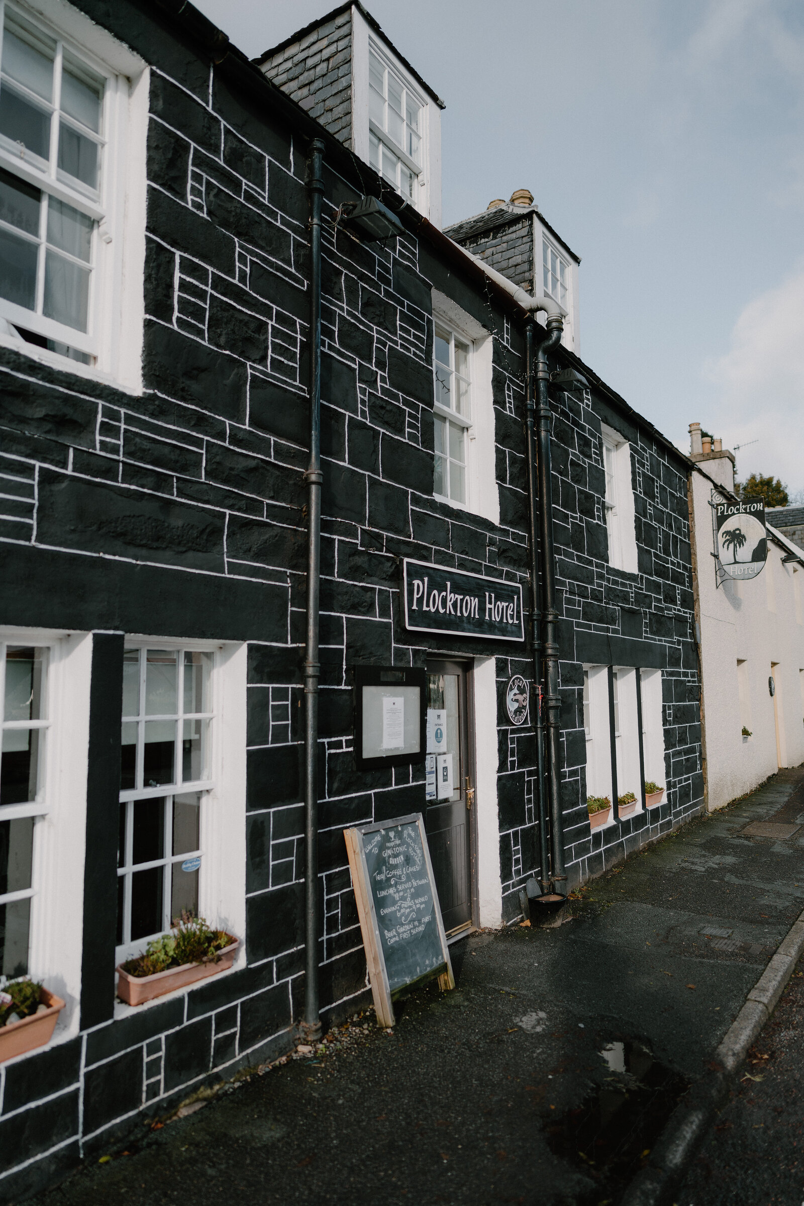 The Plockton Hotel's black-and-white harled facade lines a quiet street, a palm tree visible next door.