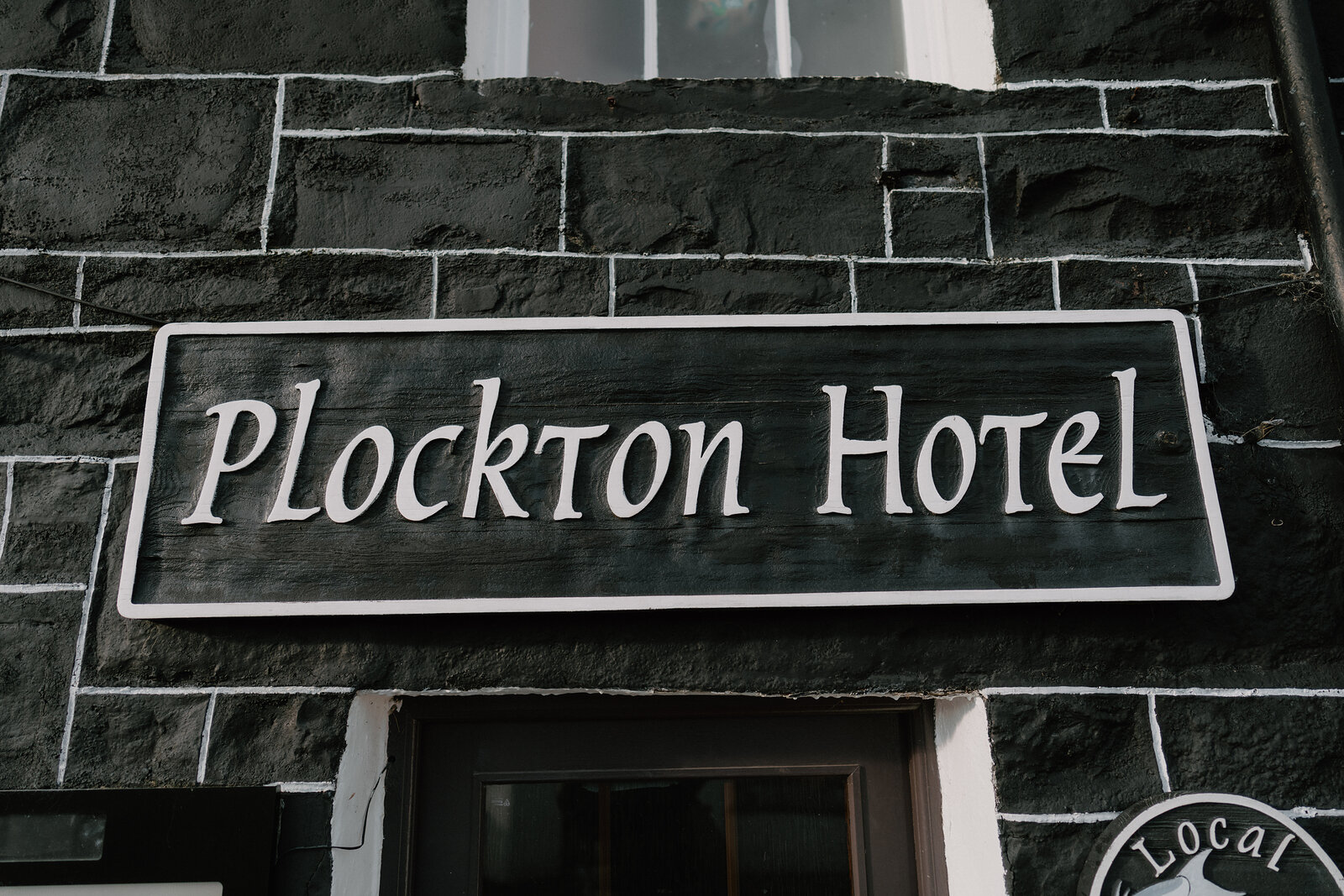 The Plockton Hotel's carved sign hangs against a black-and-white stone wall.