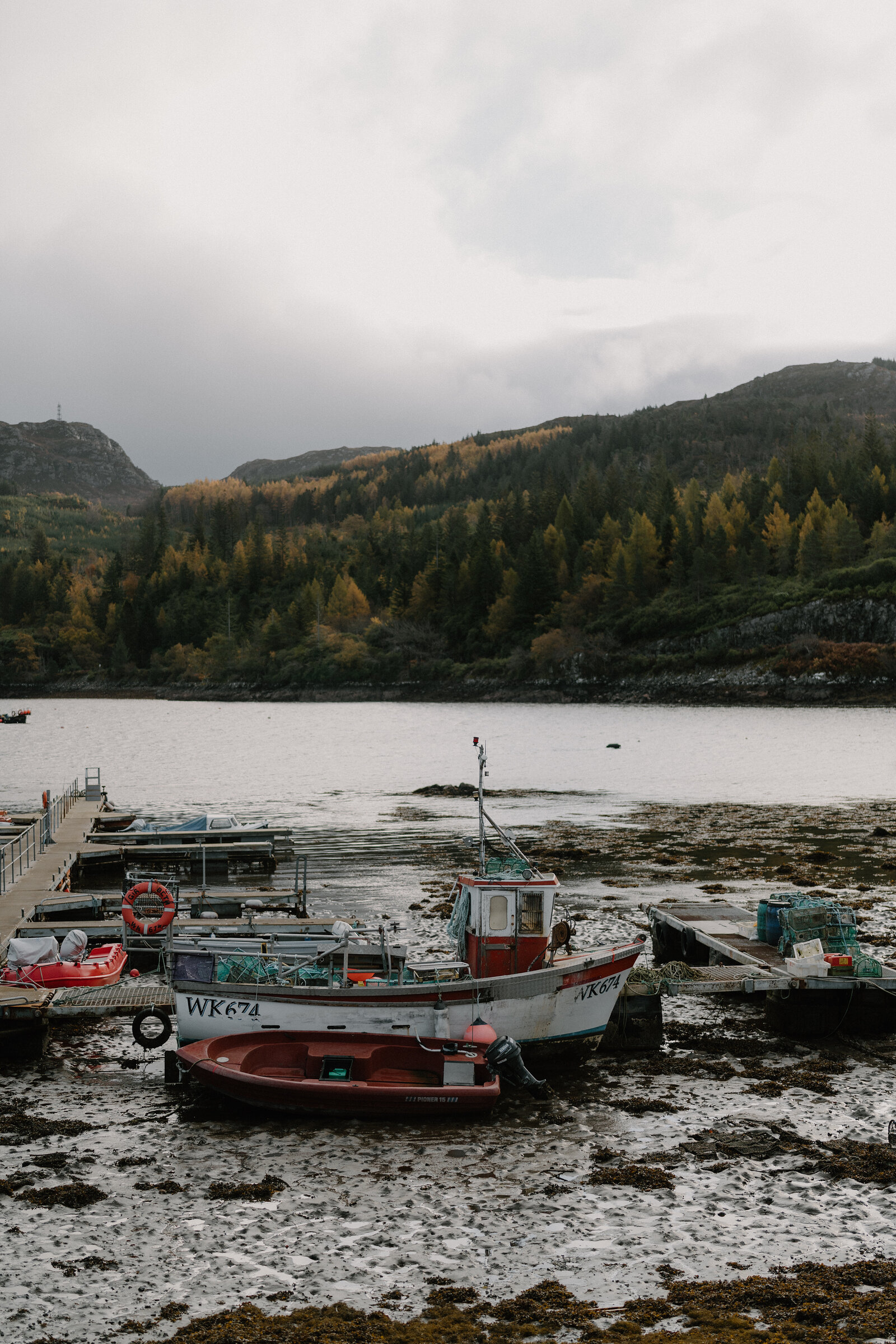 Fishing boats rest on the tideline at Plockton harbour, autumn woodland rising behind.