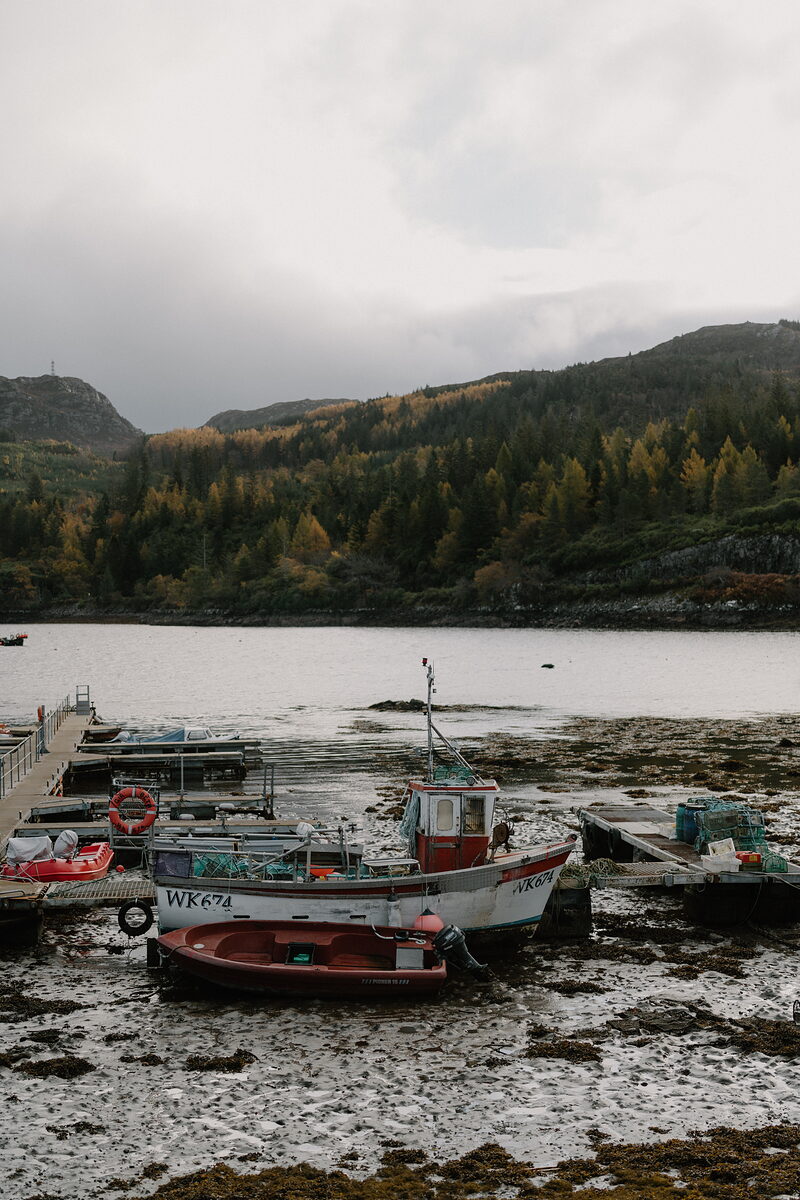Fishing boats rest on the tideline at Plockton harbour, autumn woodland rising behind.