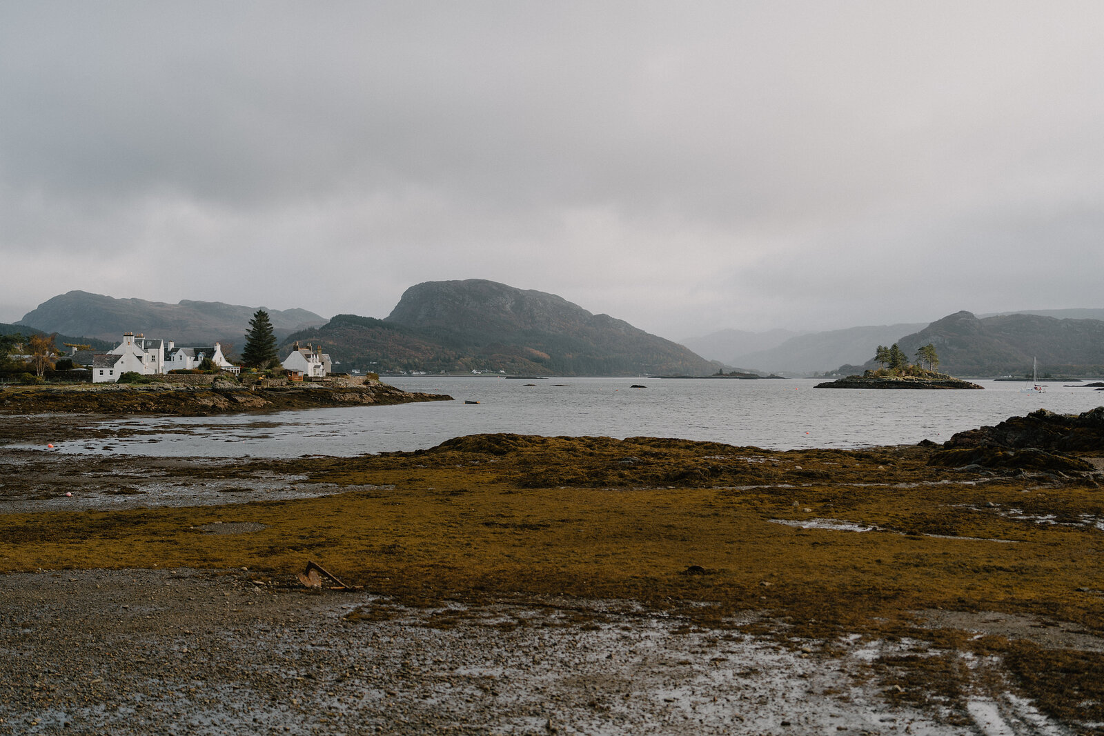 White houses line Plockton's shore below misty hills, seaweed covering the foreshore at low tide.