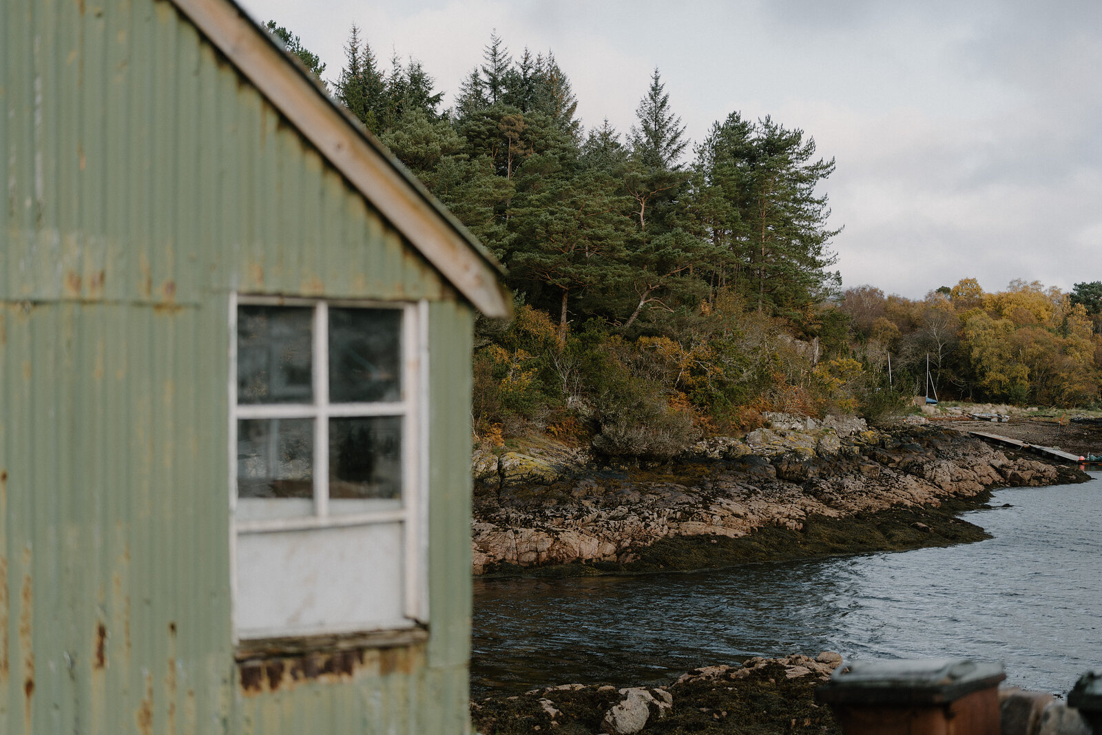 A rusted green corrugated shed overlooks a wooded, rocky shoreline in Ross and Cromarty.