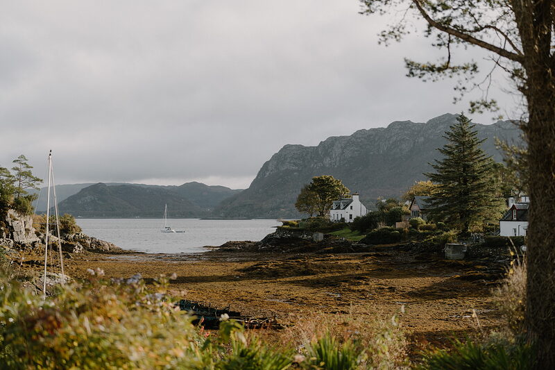A white cottage overlooks a loch where a sailboat rides at anchor, hills rising beyond.