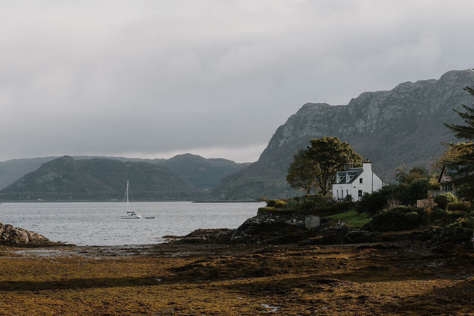 A white cottage with a lit chimney stands above a seaweed shore, a sailboat anchored offshore.