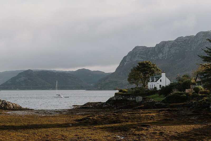A white cottage with a lit chimney stands above a seaweed shore, a sailboat anchored offshore.