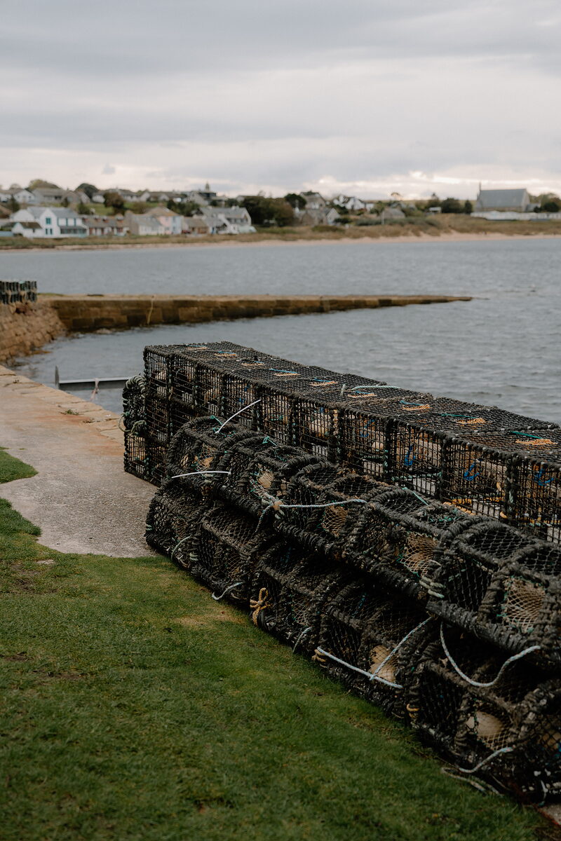 Creels stacked on a grassy bank overlook a Ross and Cromarty harbour, the village and a church spire visible across the water.