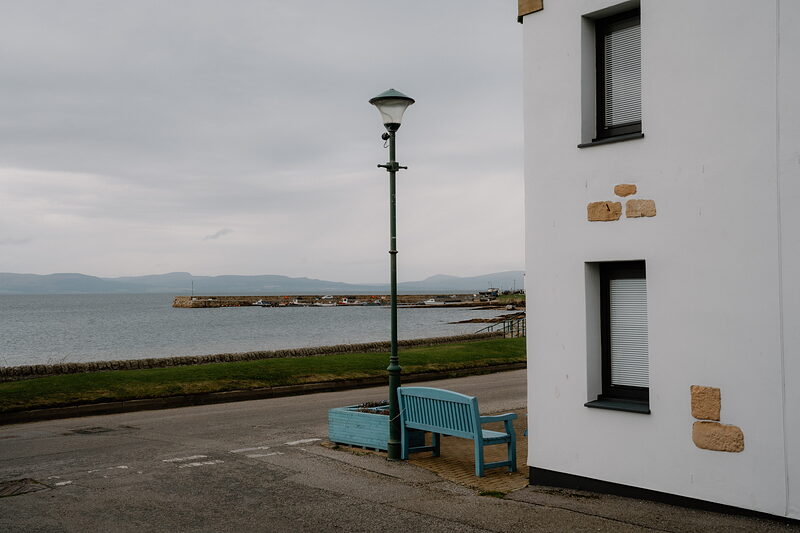 A lamppost and bench sit at a street corner above a Ross and Cromarty harbour, hills fading into haze across the water.