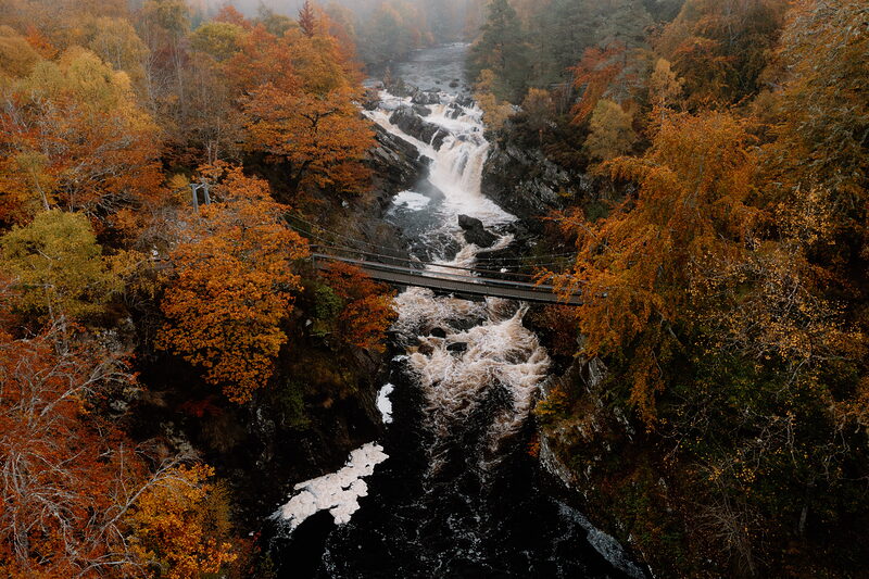 A suspension footbridge crosses a misty autumn gorge above a waterfall in Ross and Cromarty, seen from above.