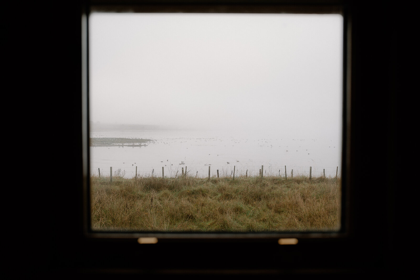 A wooden hide window frames a misty view of a fenced tidal flat in Ross and Cromarty.