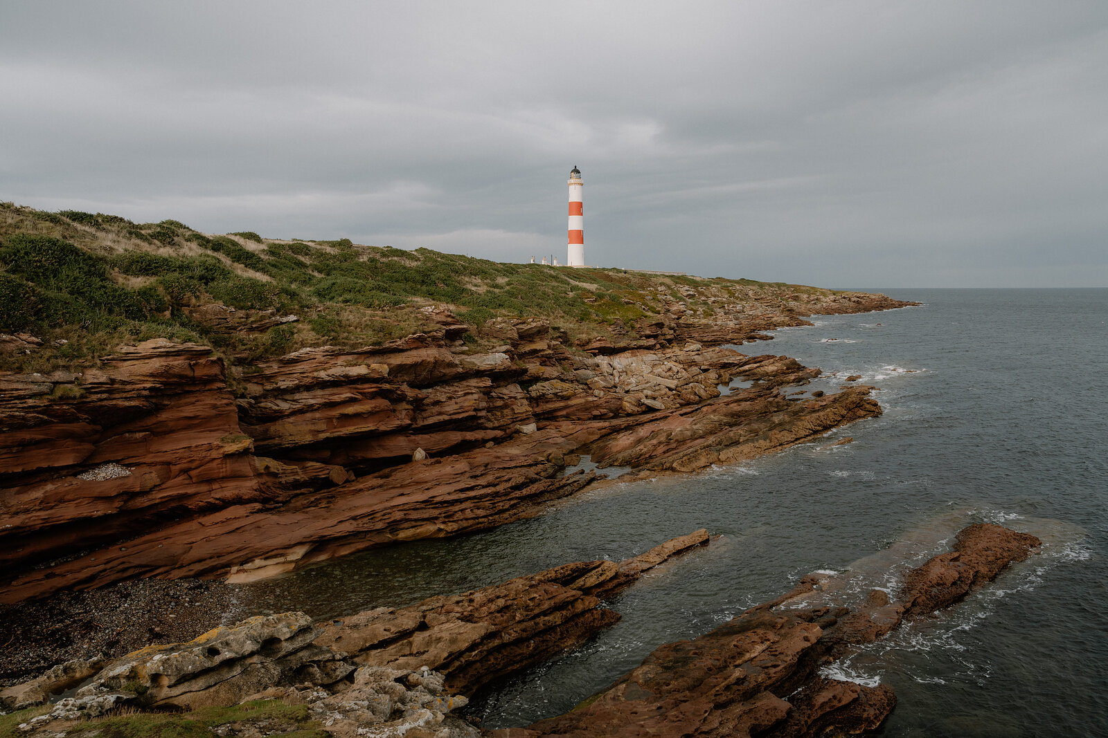 Ledges of red sandstone running out into a grey sea, the lighthouse on the green headland beyond.