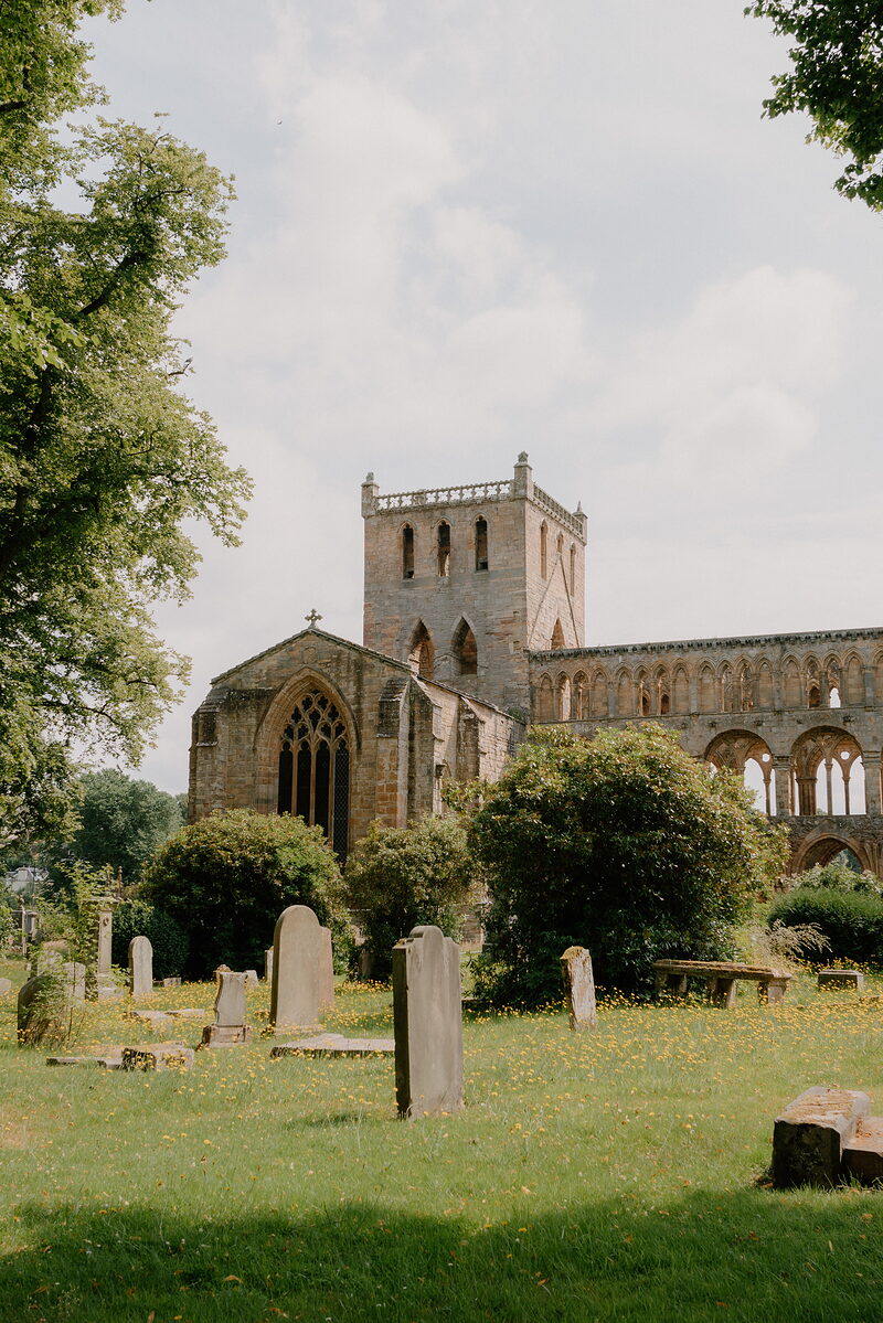 An intact abbey tower with battlements rises beyond a gothic window and shrubs, gravestones scattered across the lawn in front.