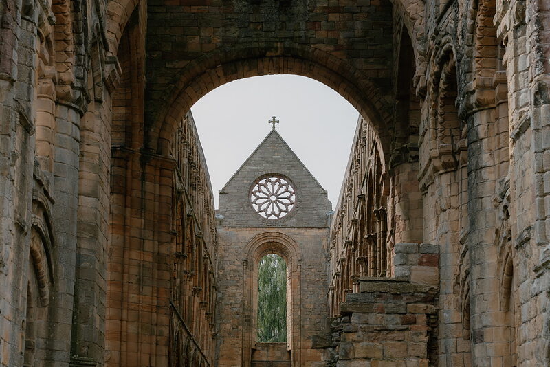 A ruined abbey's gothic arch frames a distant rose window and cross, a gravel path leading through the tiered arches.