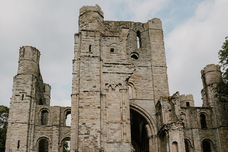 An intact abbey gatehouse tower with corner turrets rises above a pointed archway, ruined wings extending on either side.