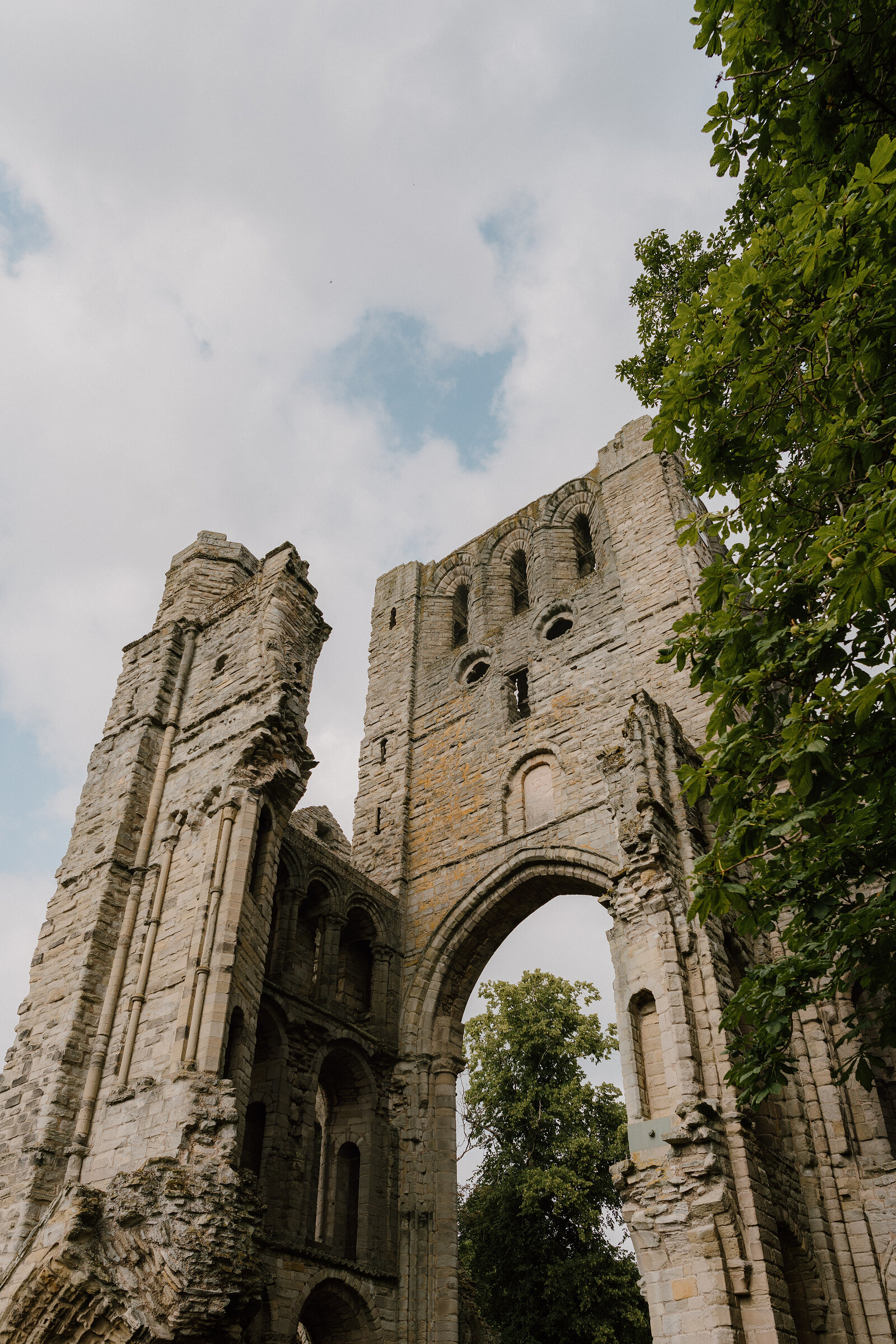 An intact abbey gatehouse with twin towers and a pointed archway is framed by overhanging leaves, ruined walls extending on either side.