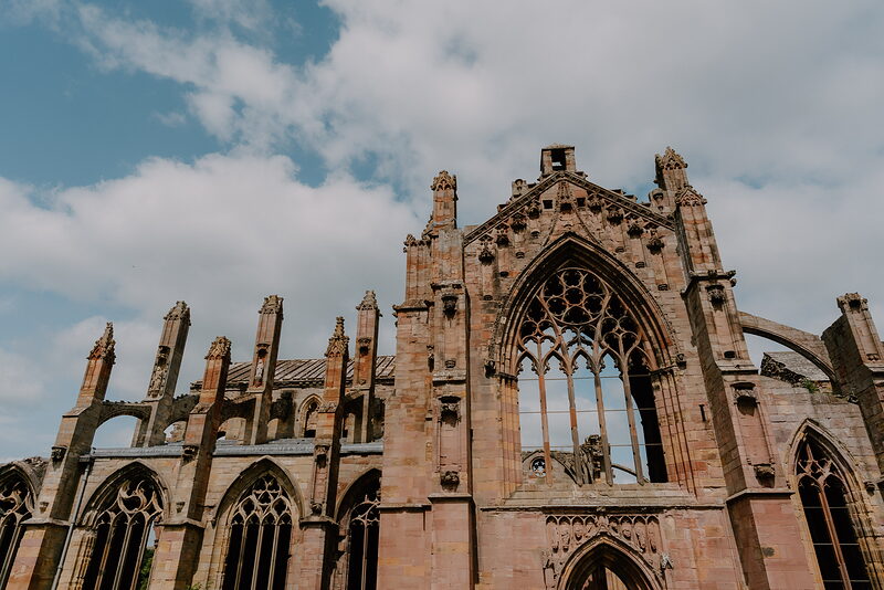 A gothic abbey facade with pinnacles, buttresses and a large traceried window rises against a partly cloudy sky.