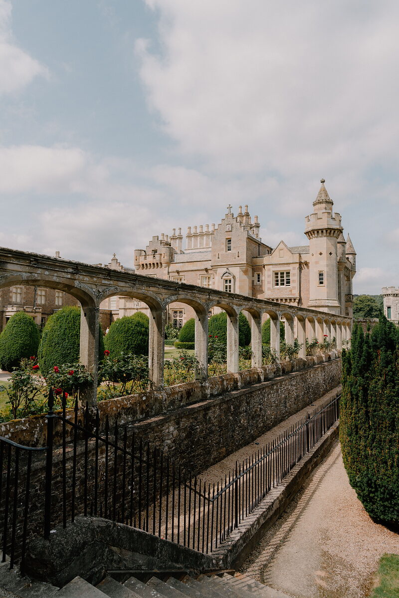 A gothic mansion with a corner turret rises beyond an arcaded stone terrace, roses climbing the wall along a rose garden below.