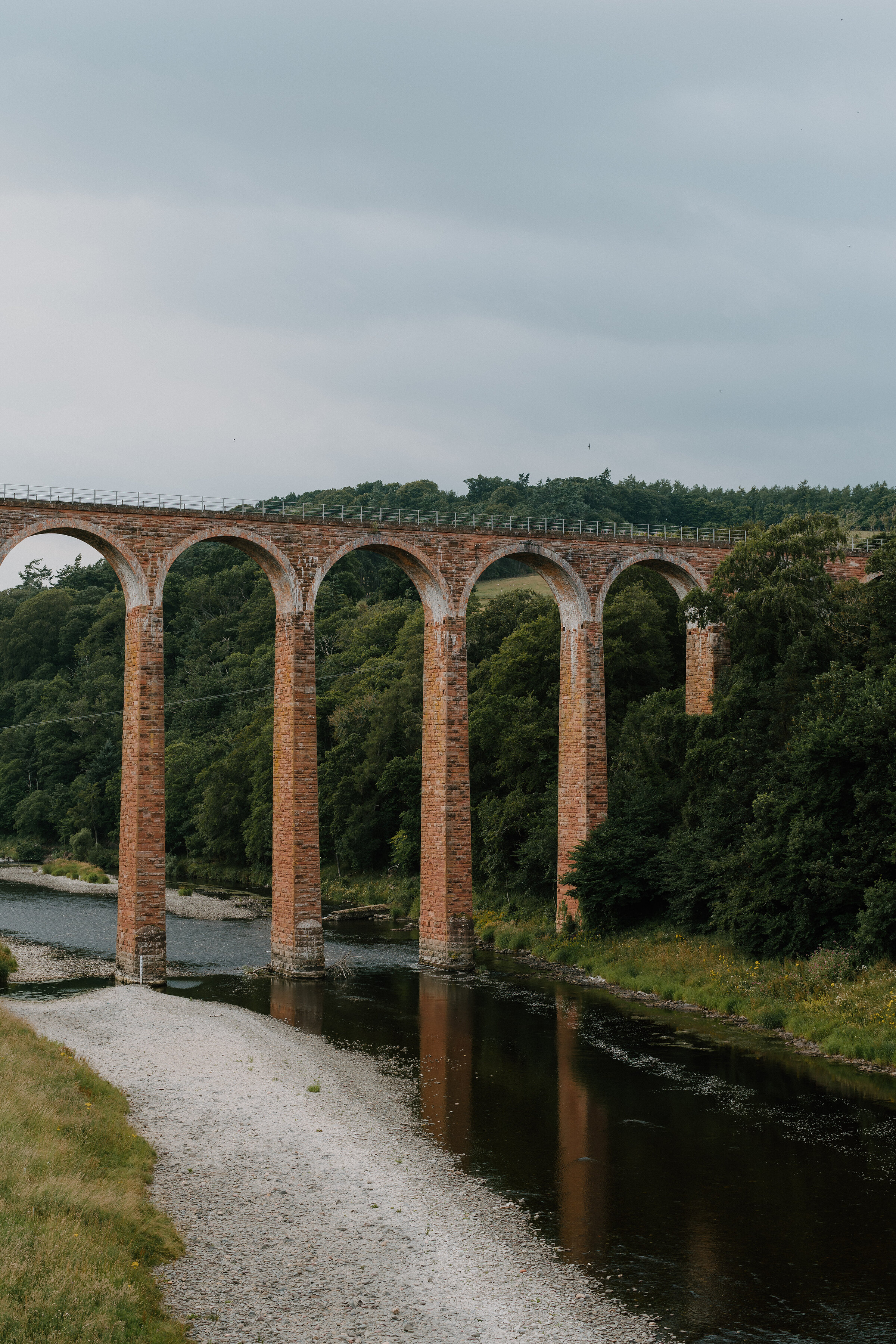 A tall brick railway viaduct with multiple arches spans a wooded river valley, a gravel path running along the bank below.
