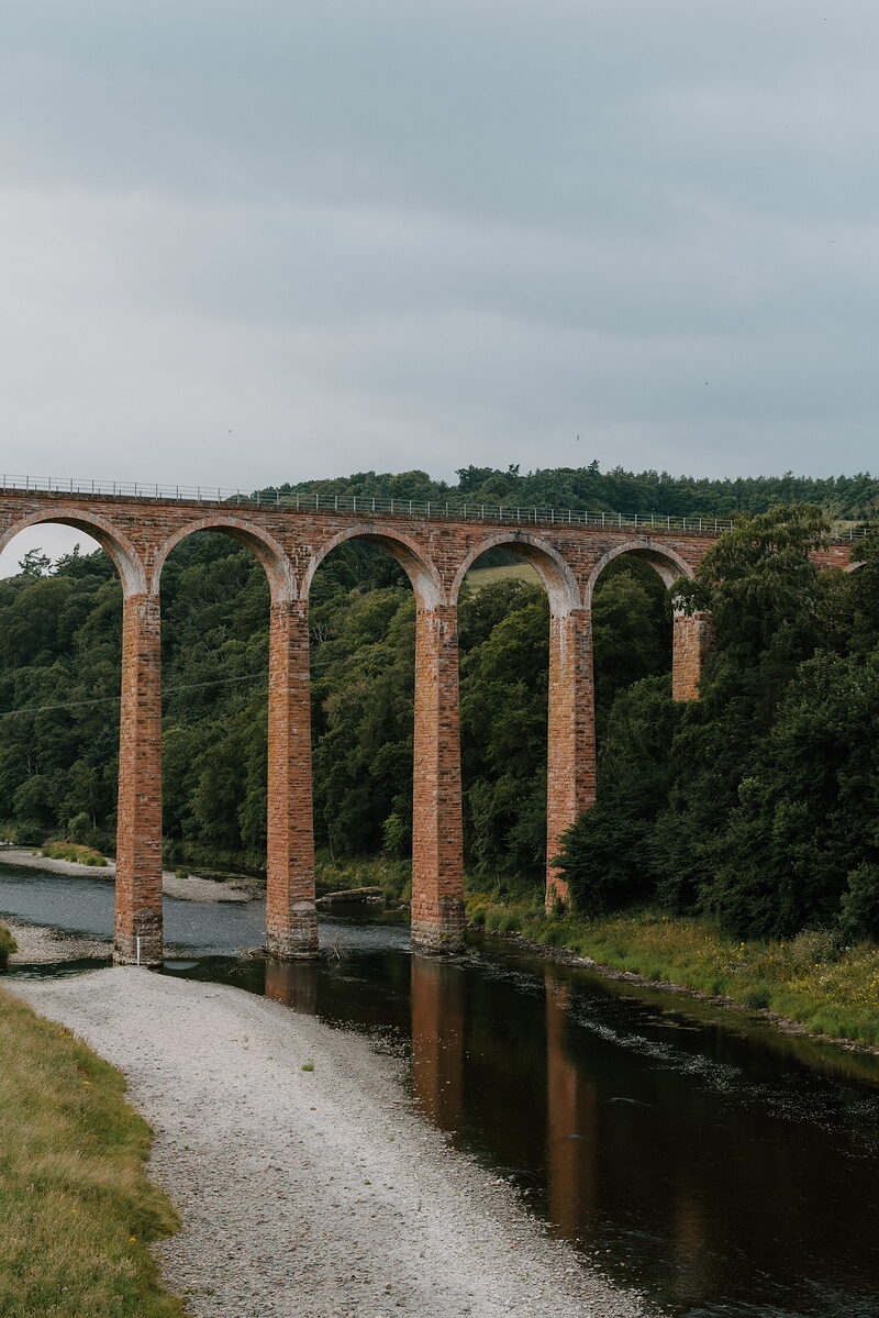 A tall brick railway viaduct with multiple arches spans a wooded river valley, a gravel path running along the bank below.