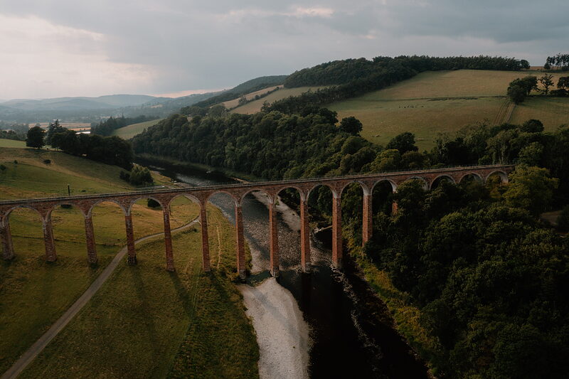 An aerial view shows a tall brick railway viaduct curving across a green valley, a river winding beneath its arches.