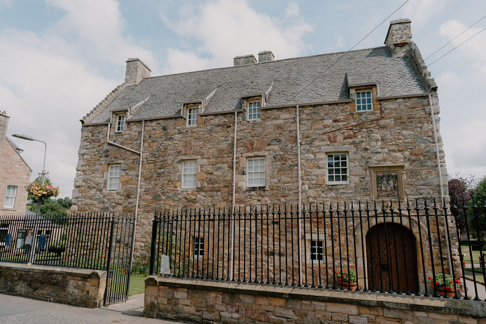 A tall stone mansion with crow-stepped gables and dormer windows stands behind iron railings, a wooden door set into an arched gateway.