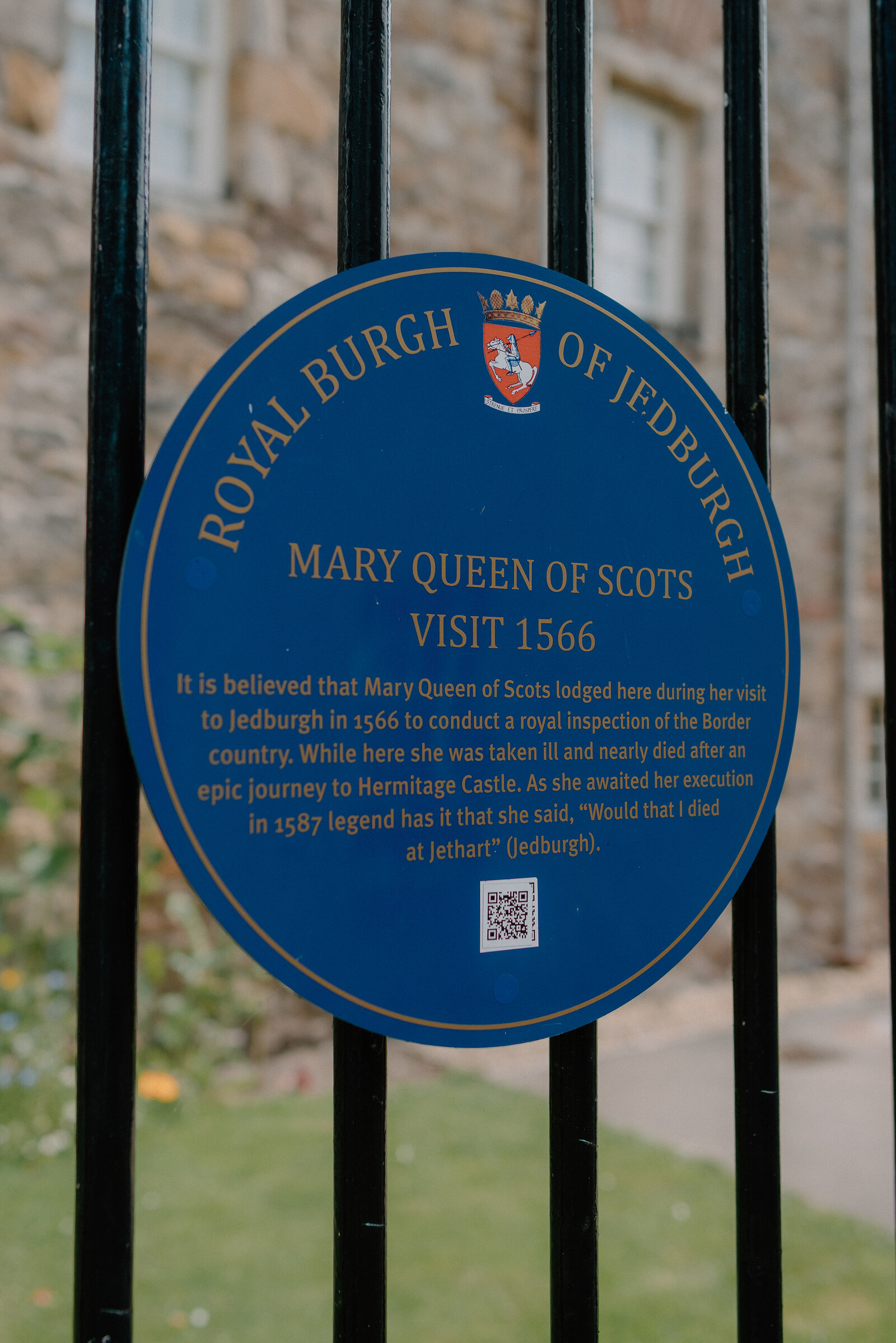 A blue plaque on iron railings reads "Royal Burgh of Jedburgh, Mary Queen of Scots Visit 1566", recounting her illness during a royal inspection of the Border country.