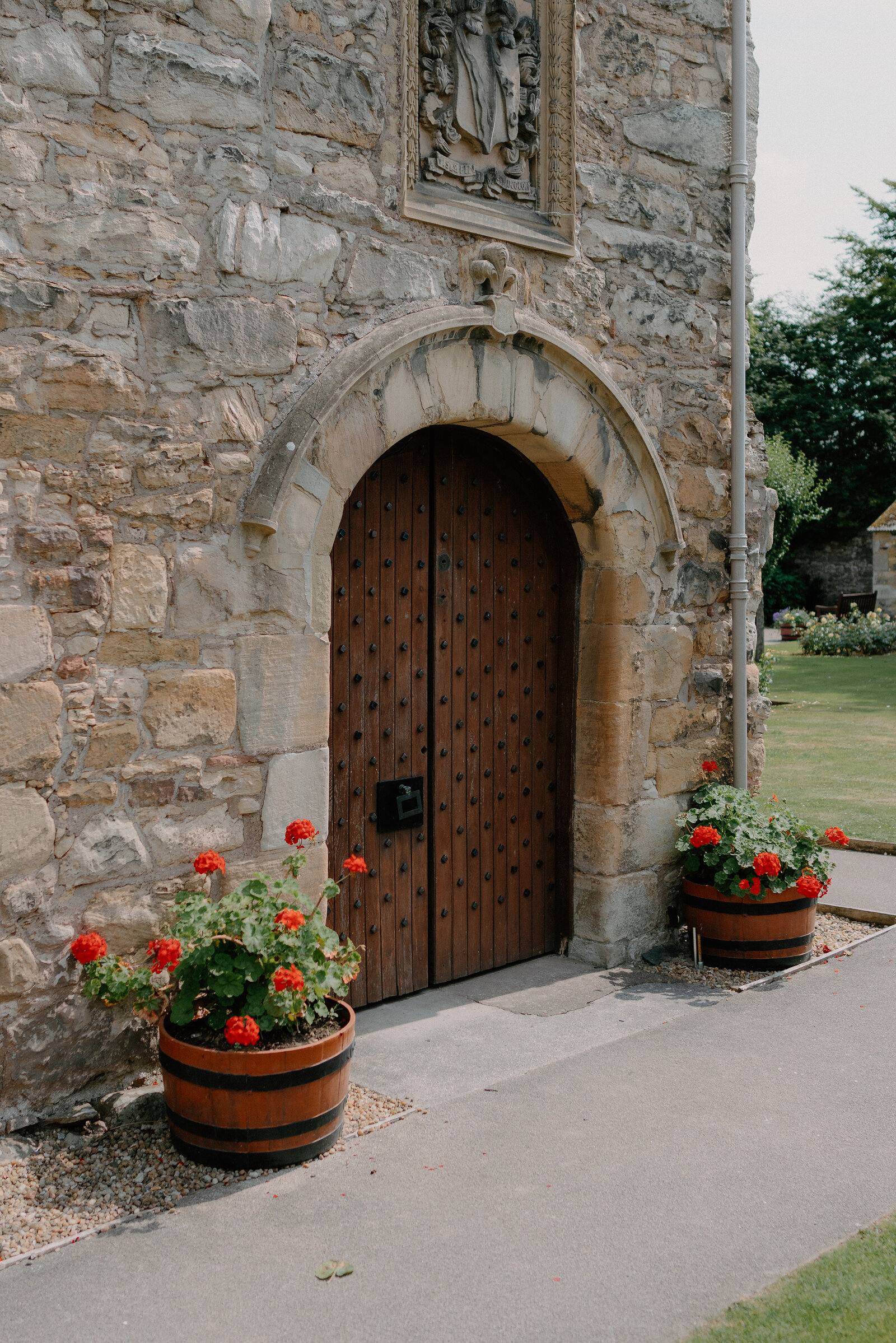 A carved stone archway frames a studded wooden door in a church wall, barrels planted with red geraniums standing on either side.