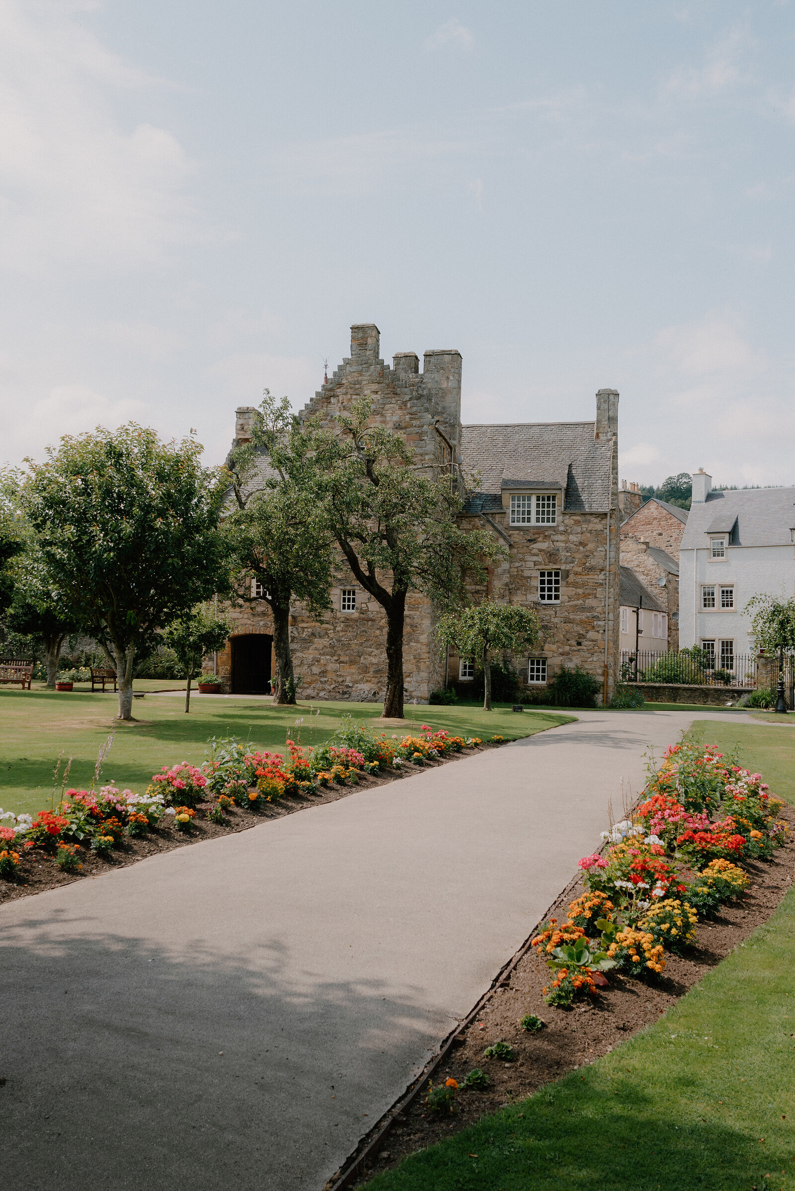 A gravel path lined with borders of red, orange and yellow flowers leads towards a turreted stone mansion, mature trees standing on the lawn beside it.