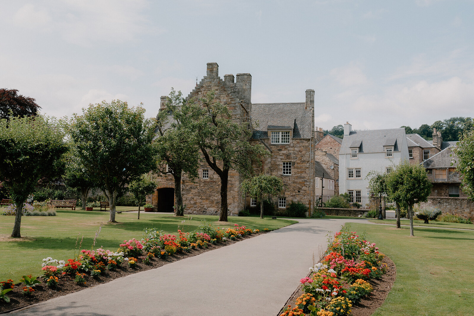 A turreted stone mansion stands beyond a lawn bordered with red and orange flowers, mature trees and a path leading towards its entrance.