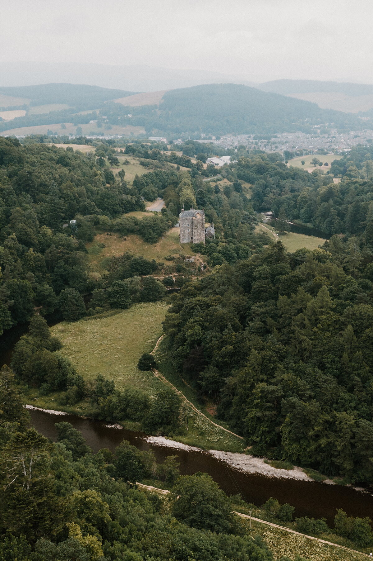 An aerial view shows a small stone tower standing in a wooded valley beside a winding river, hills and a distant town spreading beyond.