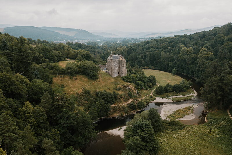 An aerial view shows a small stone tower standing on a wooded knoll above a river, hills and a distant town spreading beyond.