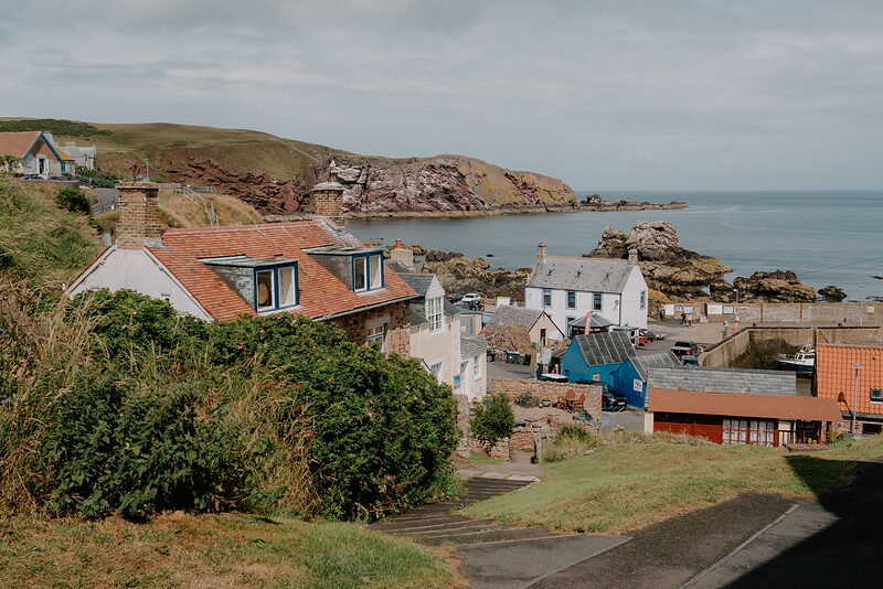 A fishing village of red-roofed cottages sits above a small bay, red sandstone cliffs and a rocky headland framing the sea beyond.