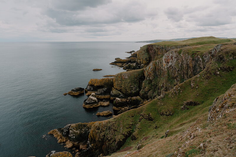 Layered green cliffs drop steeply to a calm sea beneath a cloudy sky, the coastline curving into the distance.
