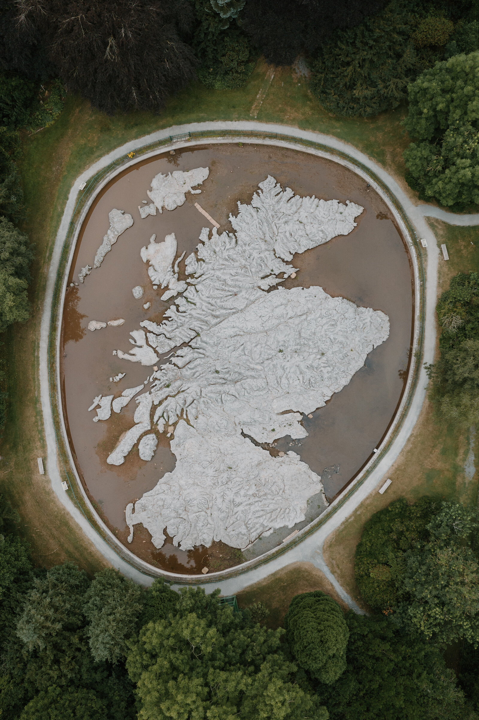 An aerial view shows a raised relief map of Scotland's coastline set in a circular pool, a path and trees ringing the installation.