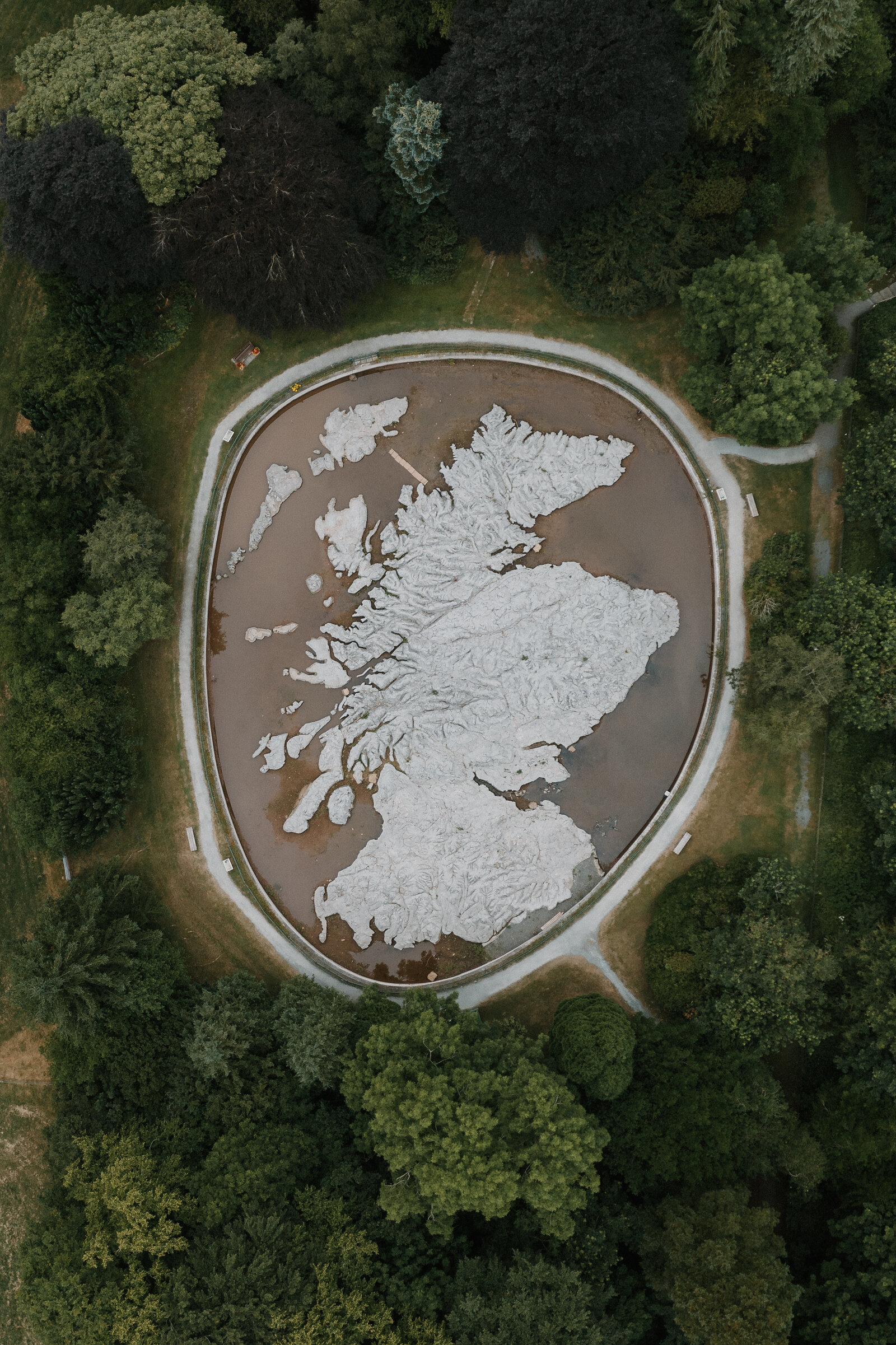 An aerial view shows a raised relief map of Scotland's coastline set in a circular pool, a path and trees ringing the installation.