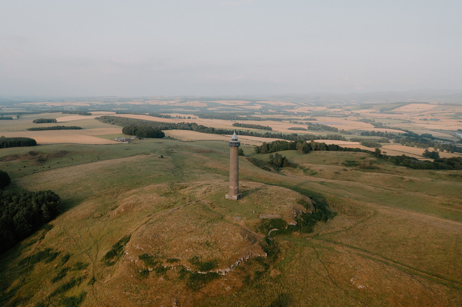 An aerial view shows a stone monument tower crowning a hilltop above patchwork farmland and scattered woodland.