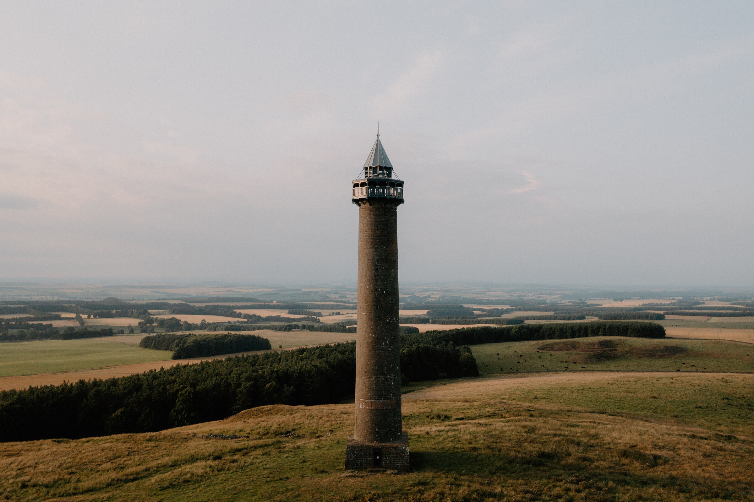A tall stone column with a lantern top stands on a hillside above patchwork farmland and woodland.