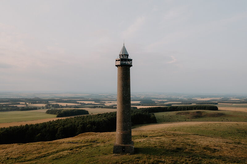A tall stone column with a lantern top stands on a hillside above patchwork farmland and woodland.