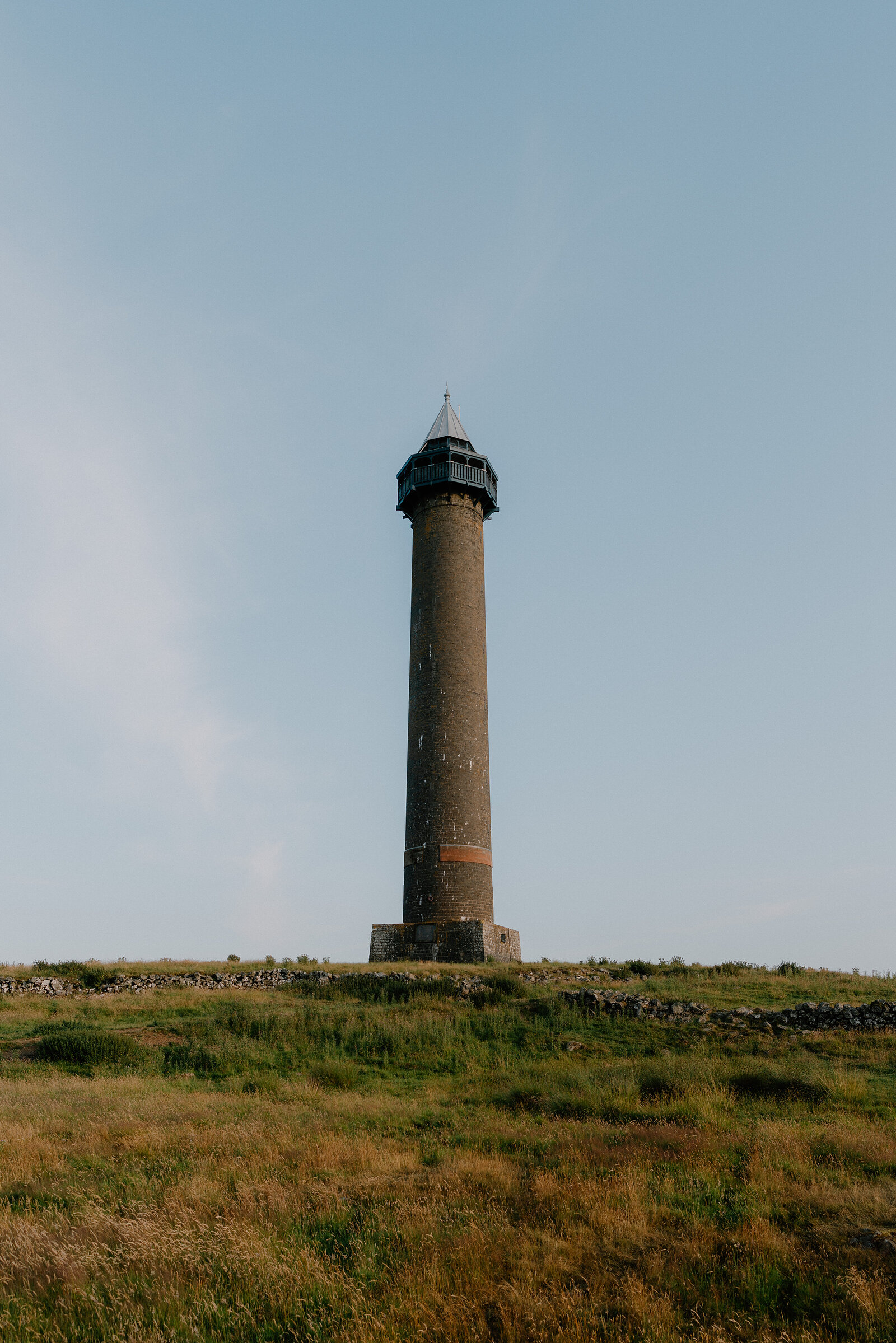 A tall stone column with a lantern top and orange stripe stands on a grassy hillside behind a dry stone wall.