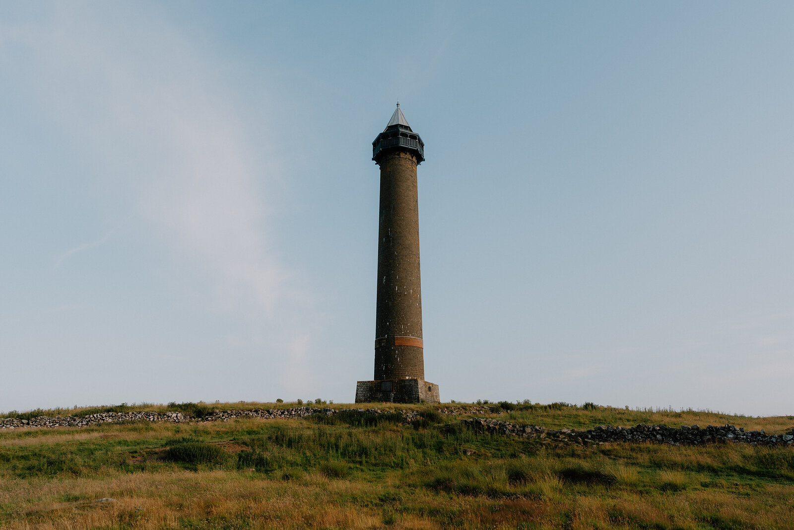 A tall stone column with a lantern top stands on a grassy hillside ringed by a dry stone wall.