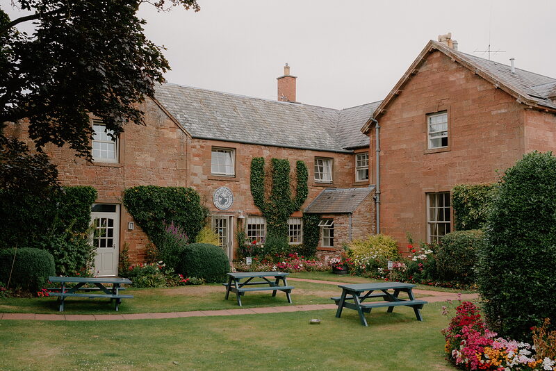 A red sandstone mansion with climbing ivy overlooks a lawn set with green picnic tables, colourful flowerbeds lining the path.
