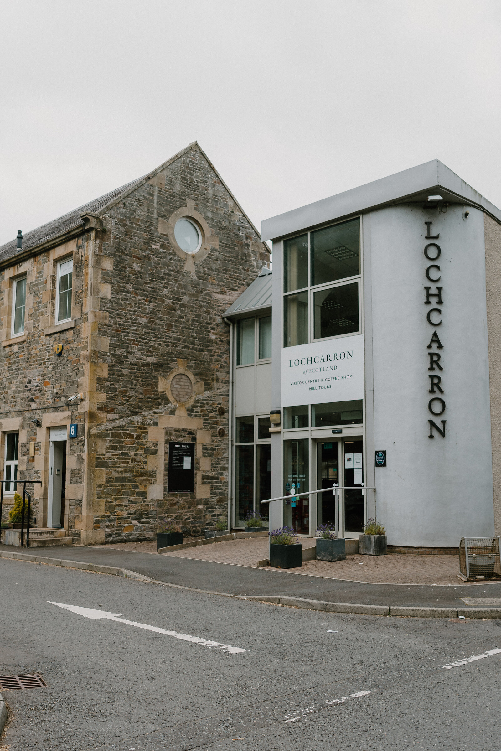 A tall grey building lettered "LOCHCARRON" stands beside an older stone building with a round window, a sign reading "Visitor Centre & Coffee Shop, Mill Tours" fixed to the glass entrance.