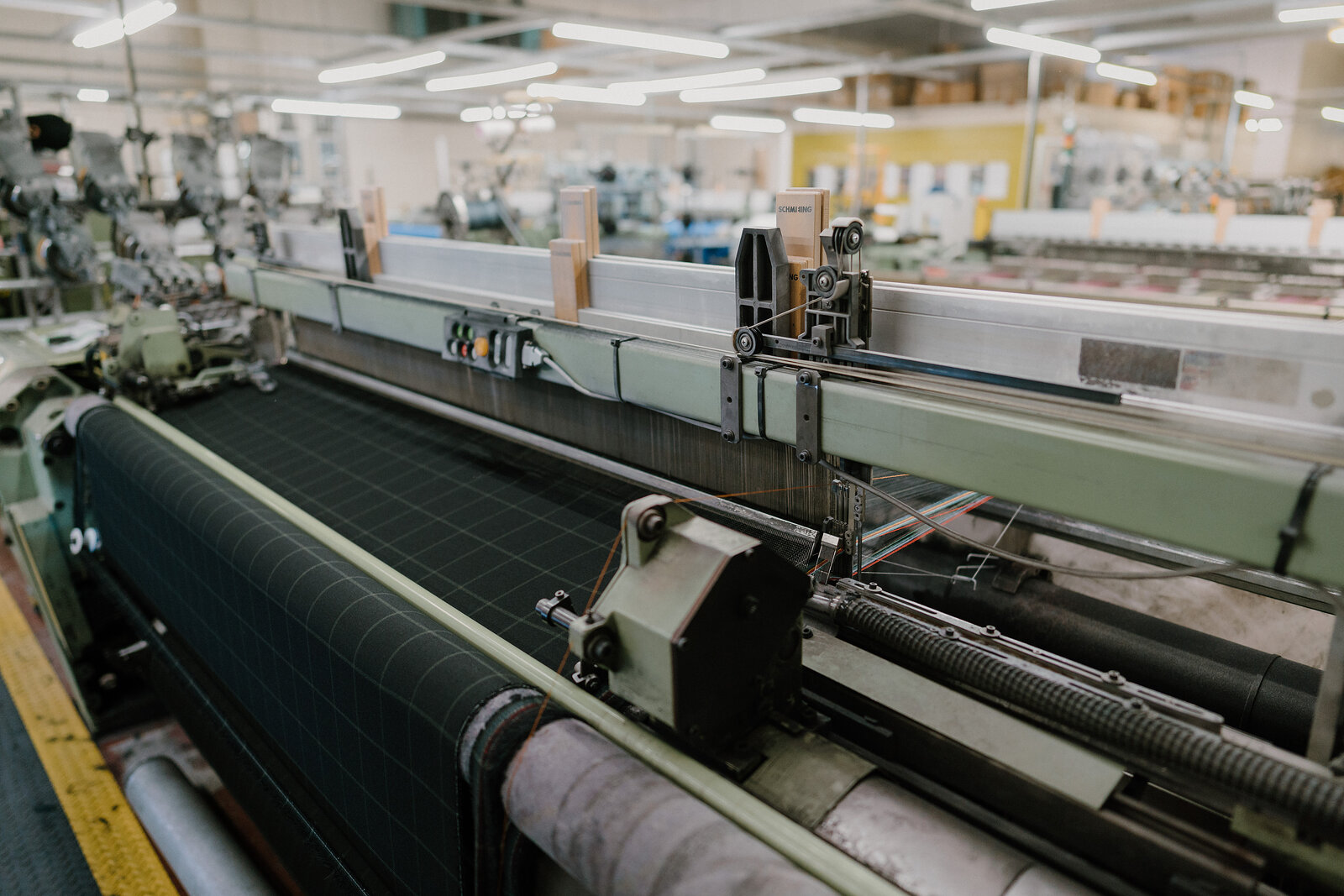 A power loom weaves dark tartan cloth on its bed, mechanical arms and bobbins visible above the woven fabric in a mill workshop.