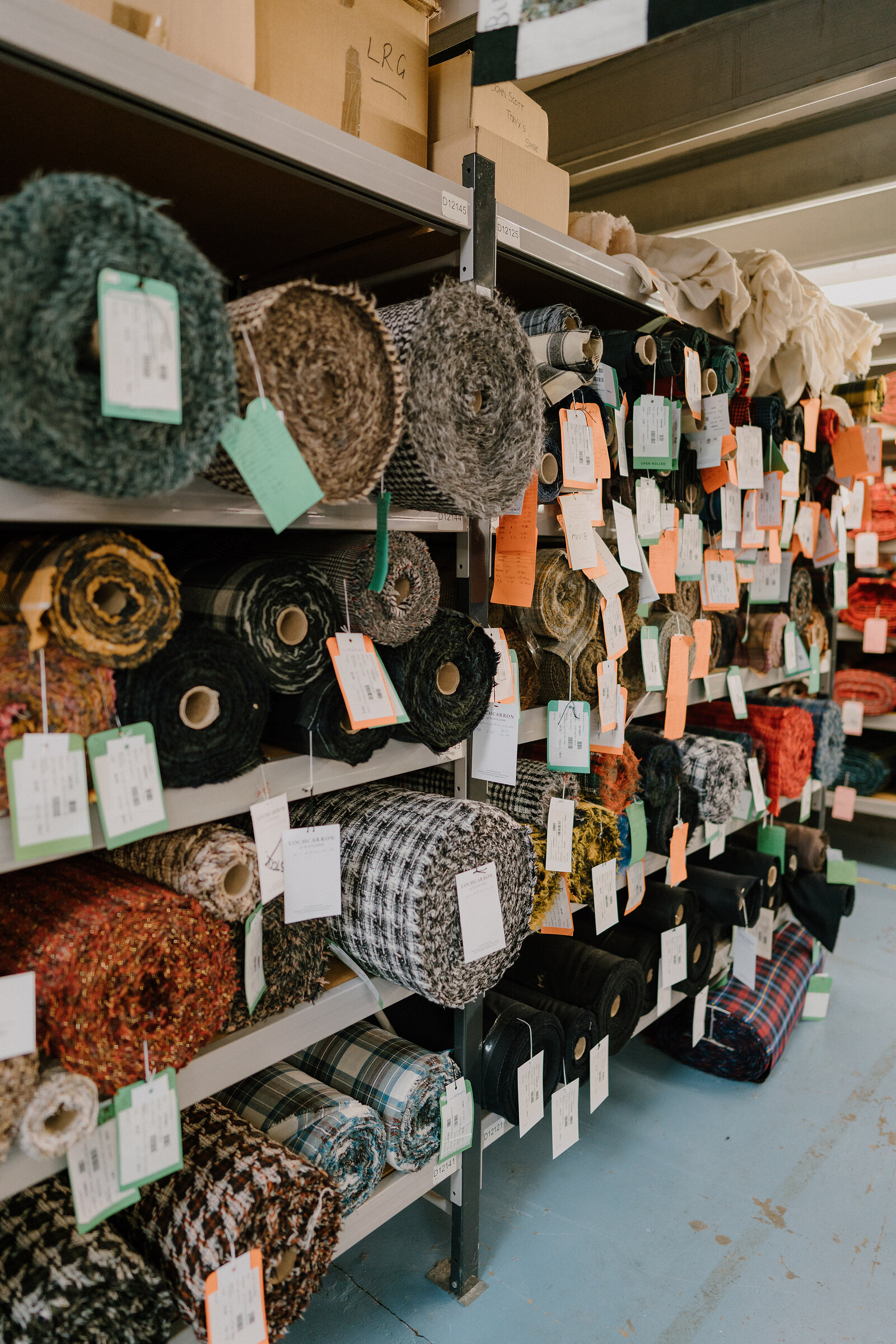 Rolls of tweed and tartan fabric in tweed, check and floral patterns stand on shelving, price tags attached to each roll.