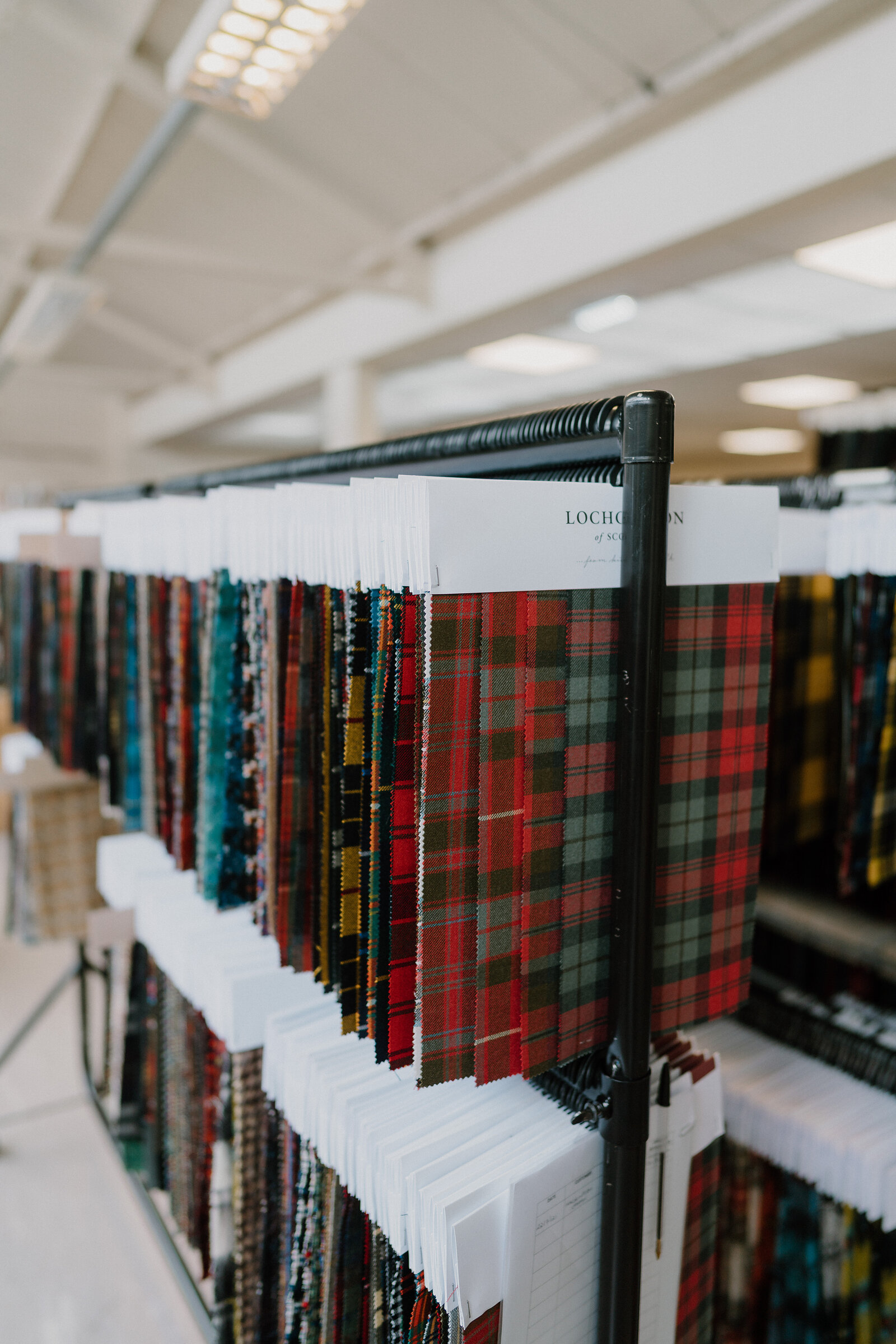 Rows of tartan fabric swatches hang from a rack labelled "Lochcarron of Scotland", red, green and yellow checks among the patterns.