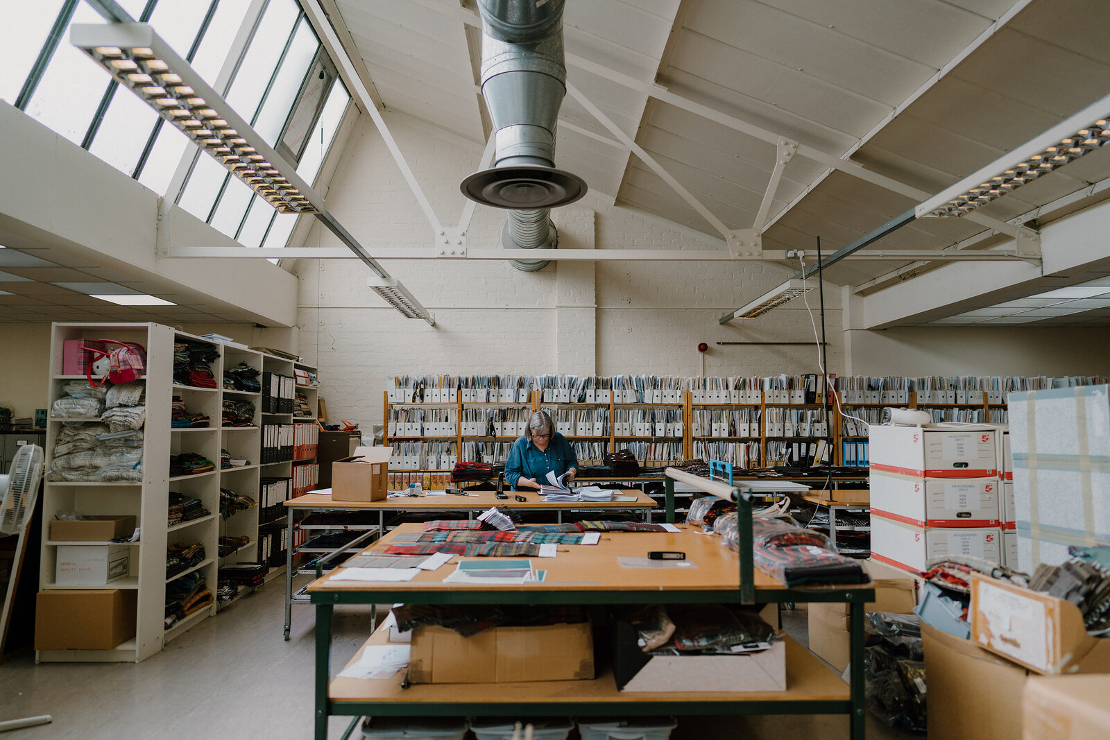 A worker sits at a table cutting fabric swatches in a bright workroom, shelving stacked with sample cards lining the wall behind.