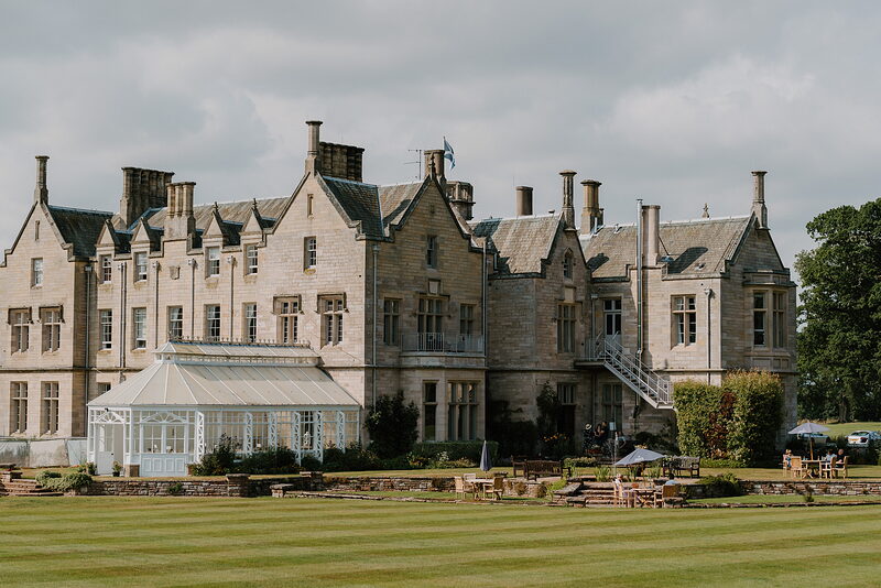 A large stone mansion with tall chimneys and gabled dormers stands behind a lawn, a glazed conservatory attached at one end and outdoor seating set on a terrace.