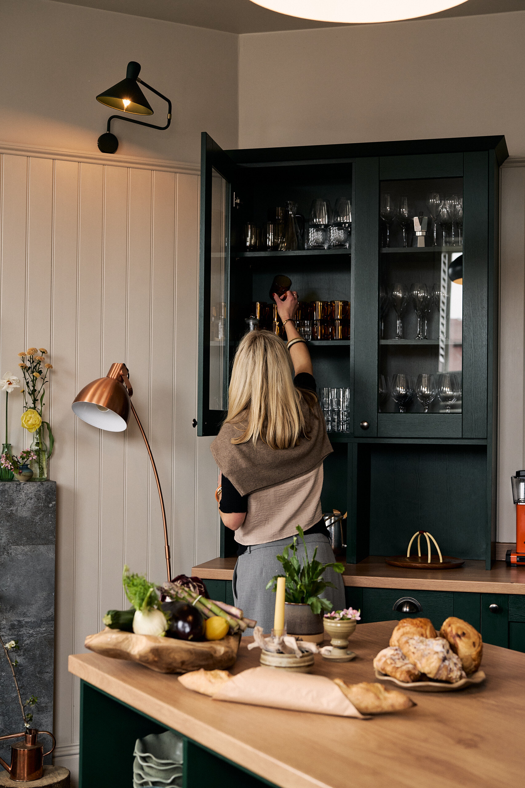 A woman in a knitted wrap reaches into an open dark green glass-fronted dresser stocked with wine glasses, a copper lamp and pastries on a wooden island in front of her.