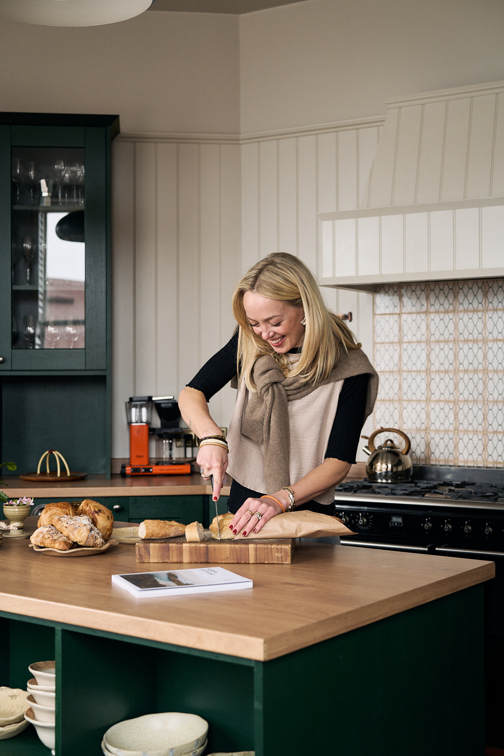 A woman in a knitted vest smiles while slicing bread on a wooden board at a kitchen island, a magazine resting on the counter beside a plate of pastries.