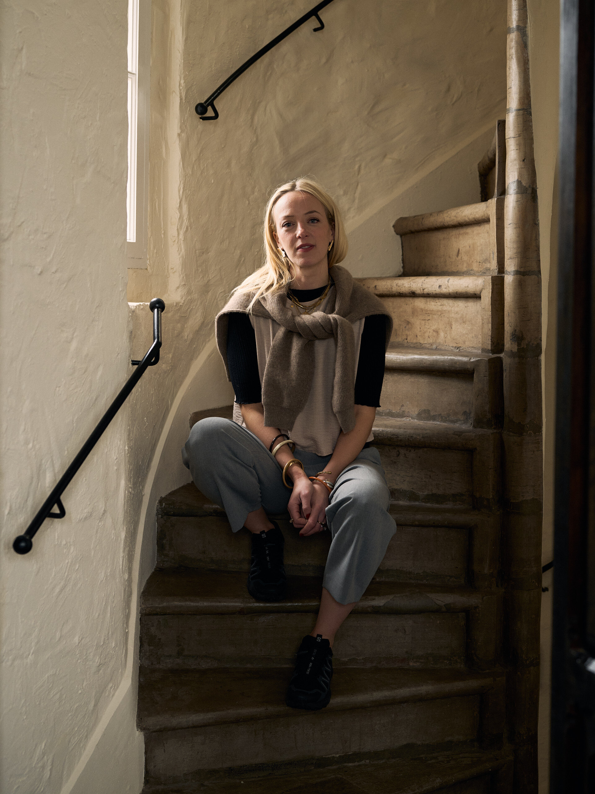 A woman in a knitted vest and layered jewellery sits looking up from a stone spiral staircase, an iron handrail curving up the wall beside her.